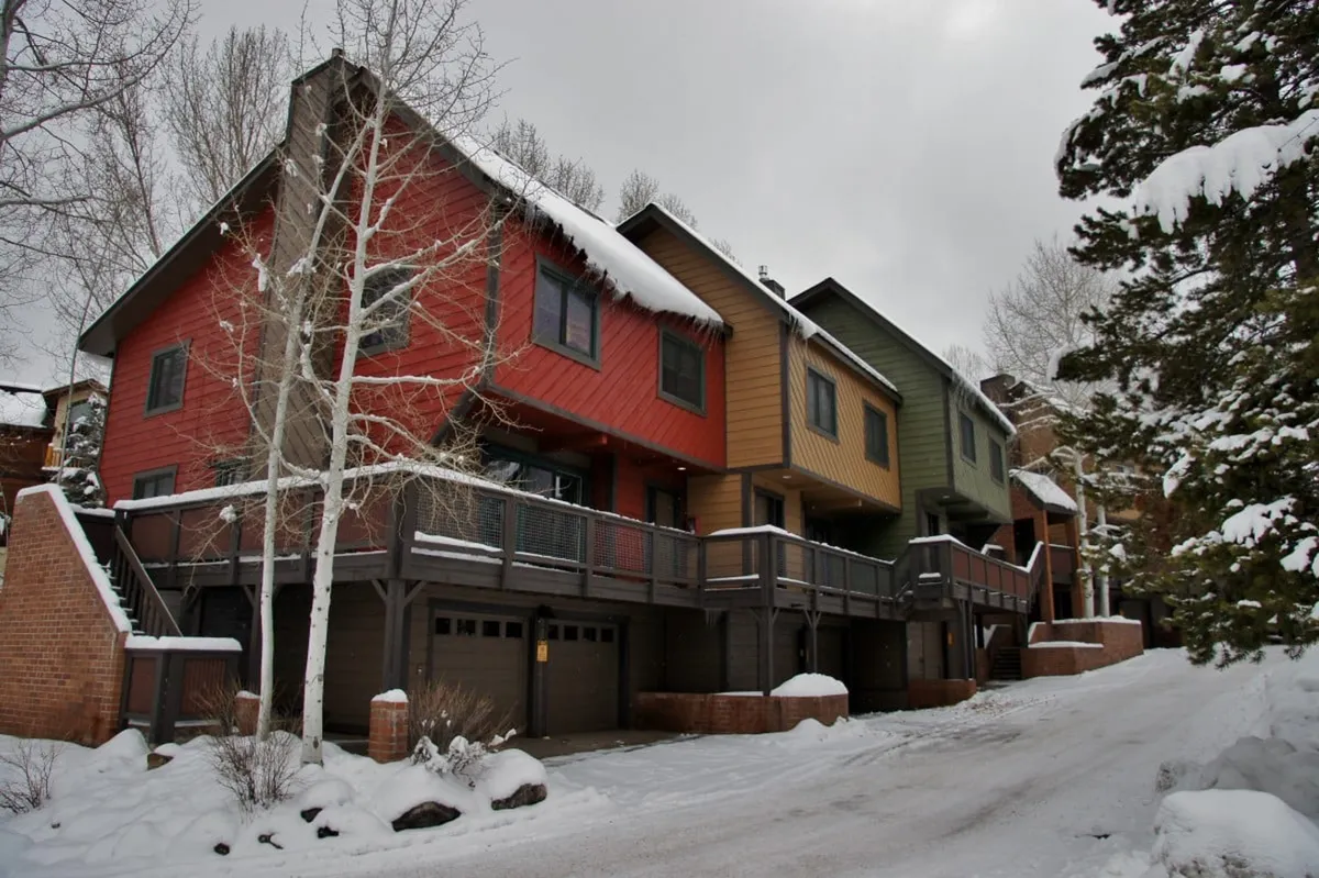 A charming winter exterior view of several colorful townhomes with snow-covered roofs nestled among tall trees. The scene suggests a cozy mountain environment ideal for ski vacations, with easy access to nearby slopes and après-ski relaxation.