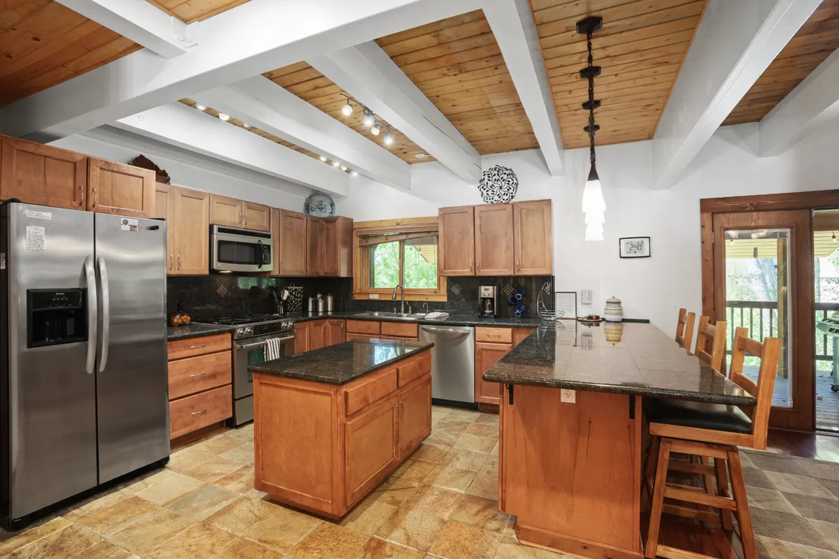 A modern kitchen with a central granite countertop island, stainless steel appliances, and wooden cabinetry. The kitchen is open to the rest of the living area and is bathed in sunlight from expansive windows, making it ideal for both casual brunches and festive gatherings.