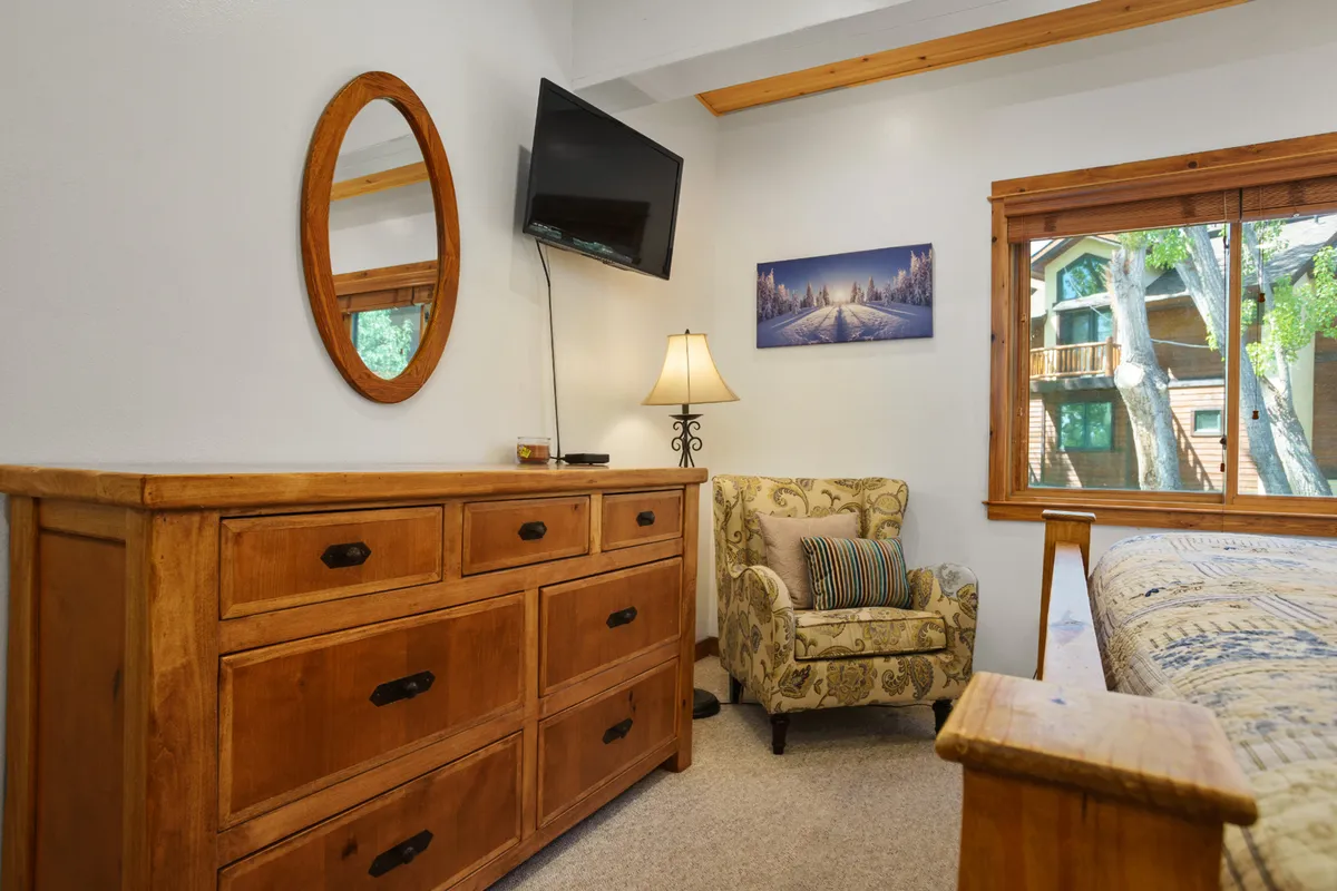 The image features a cozy reading nook with a wooden dresser, an armchair placed by a window offering a view of nature, wall art, and a lamp. The window view establishes a connection to the natural surroundings and suggests a tranquil, comfortable setting for long-term stays or remote work.