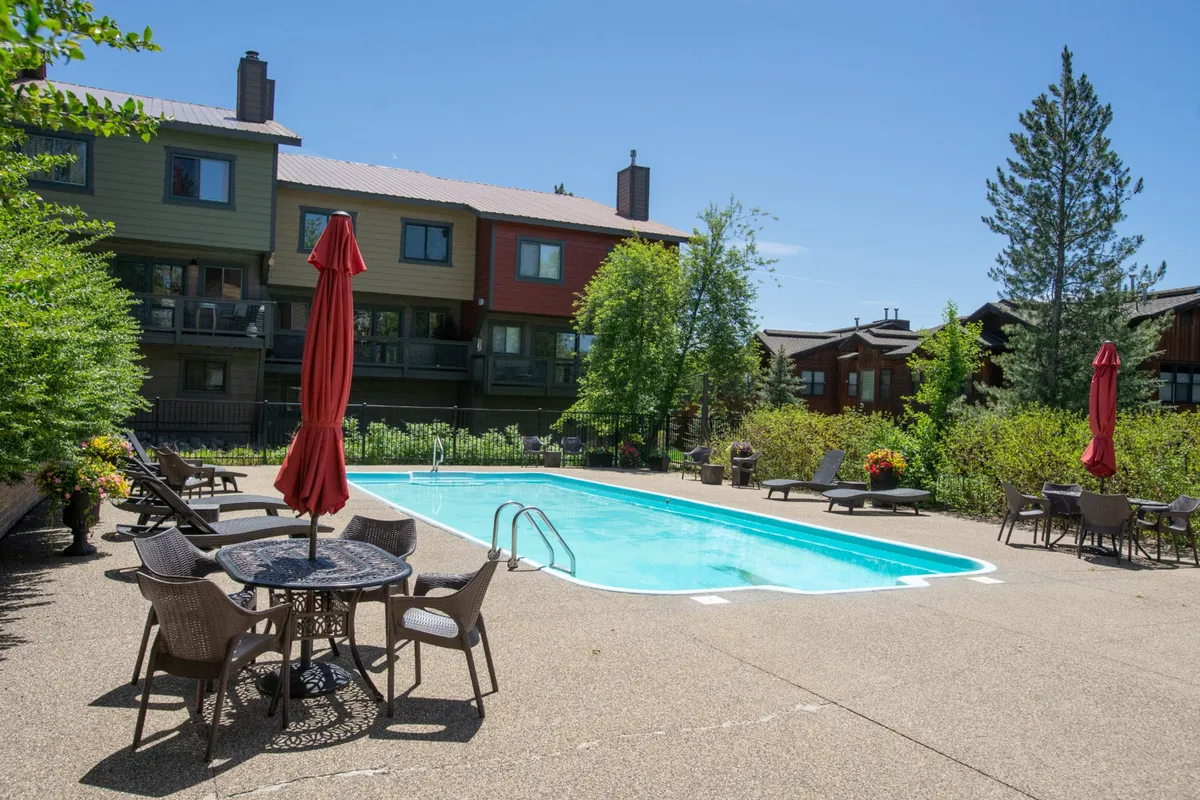 An inviting exterior pool area with a sparkling swimming pool, vibrant red umbrellas, outdoor seating, and lush greenery. The scene conveys a cheerful, social atmosphere ideal for summer sunbathing and poolside gatherings.