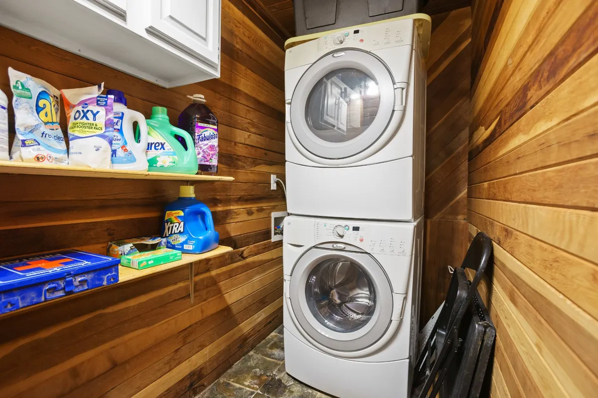 A practical, well-lit laundry area featuring a stacked washer and dryer, as well as organized shelving stocked with cleaning supplies. The space is clean, tidy, and clearly designed for ease of use, making laundry tasks simple and convenient for guests.